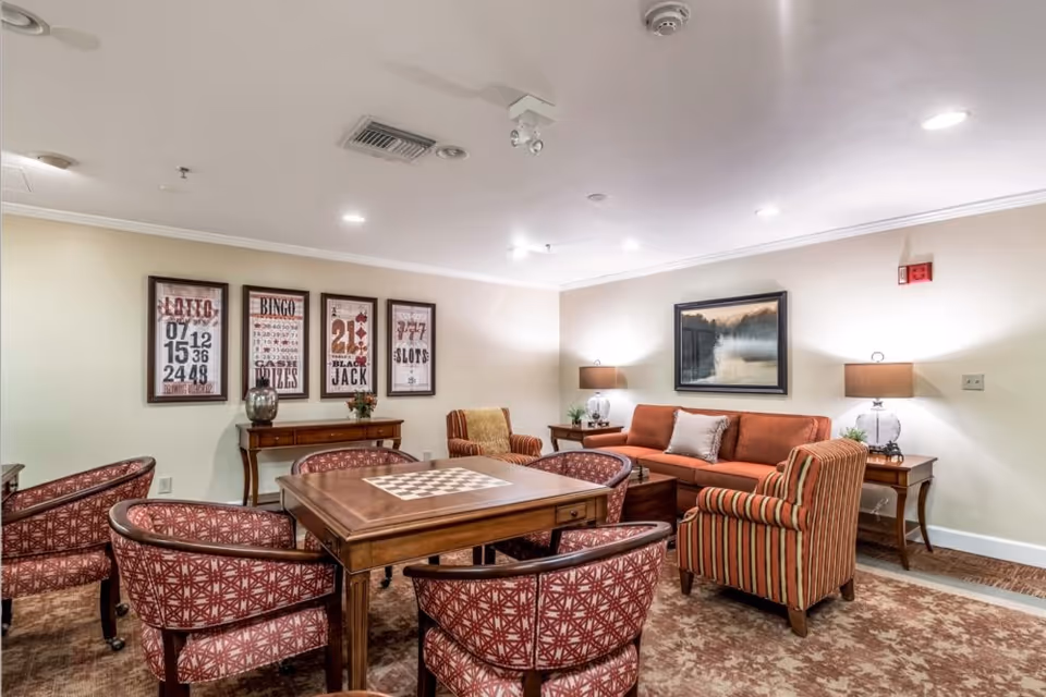 A cozy common area in a senior living facility featuring a wooden table with a built-in chessboard surrounded by six patterned armchairs. Behind the table is a wooden console table with decorative items and four framed posters on the wall advertising lotto, bingo, blackjack, and slots. To the right, there is an orange sofa with a striped armchair, two side tables with lamps, and a framed landscape painting on the wall. The room has recessed ceiling lights and a carpeted floor.