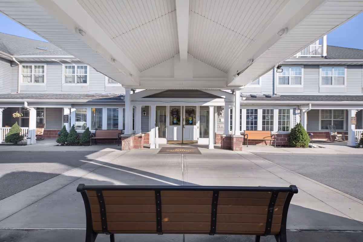 Covered entrance area of a senior living facility with a bench in the foreground, two benches on either side near the building, and a double-door entrance under a peaked roof. The building is light gray with white trim and multiple windows.