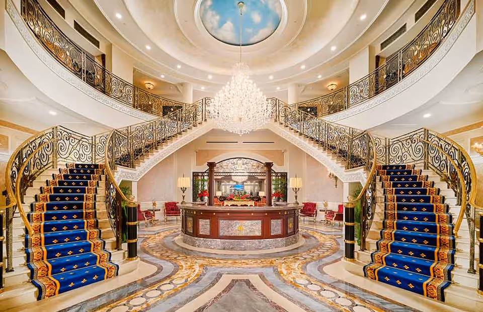 Luxurious grand lobby with a double staircase featuring ornate black and gold railings and blue carpet runners with gold patterns. A large crystal chandelier hangs from a ceiling dome painted with a sky and clouds motif. The circular reception desk is in the center, surrounded by elegant marble flooring with intricate designs. The space is well-lit with wall sconces and table lamps, and there are red upholstered chairs along the walls.