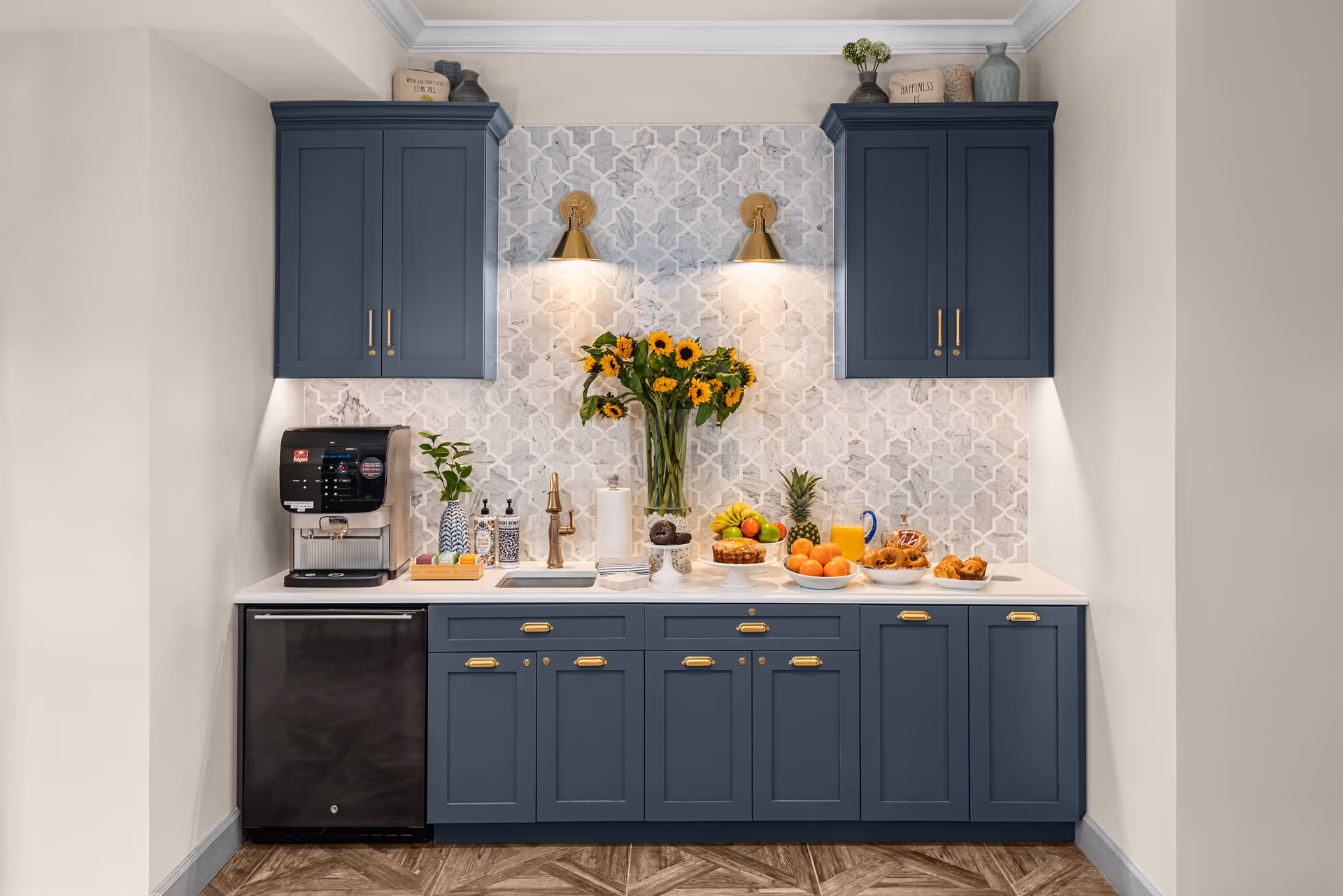 A kitchen area with blue cabinets and a white countertop. There is a coffee machine on the left, a vase with sunflowers in the center, and plates of fruit and pastries on the right. The backsplash features a patterned tile design, and two brass wall sconces provide lighting above the countertop.
