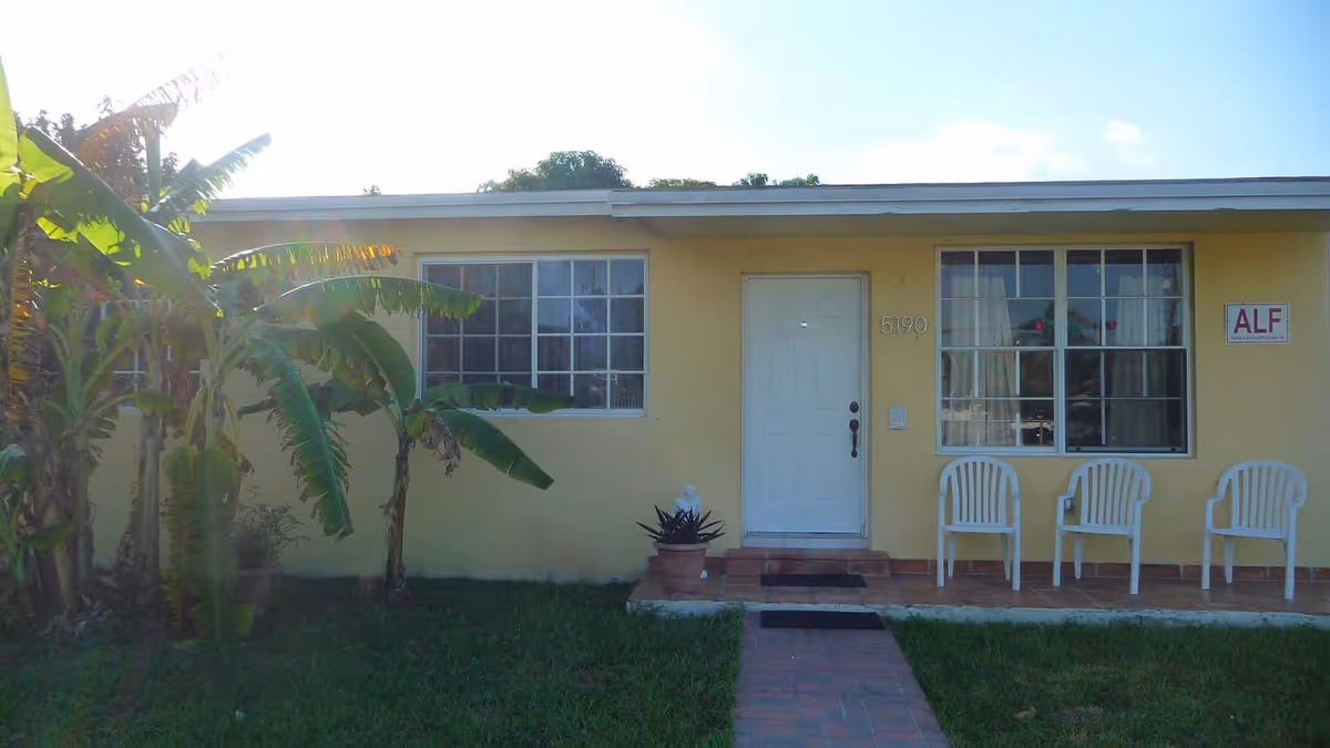Front of a single-story yellow home with a white door, two windows, three white plastic chairs on the porch and an 'ALF' sign.