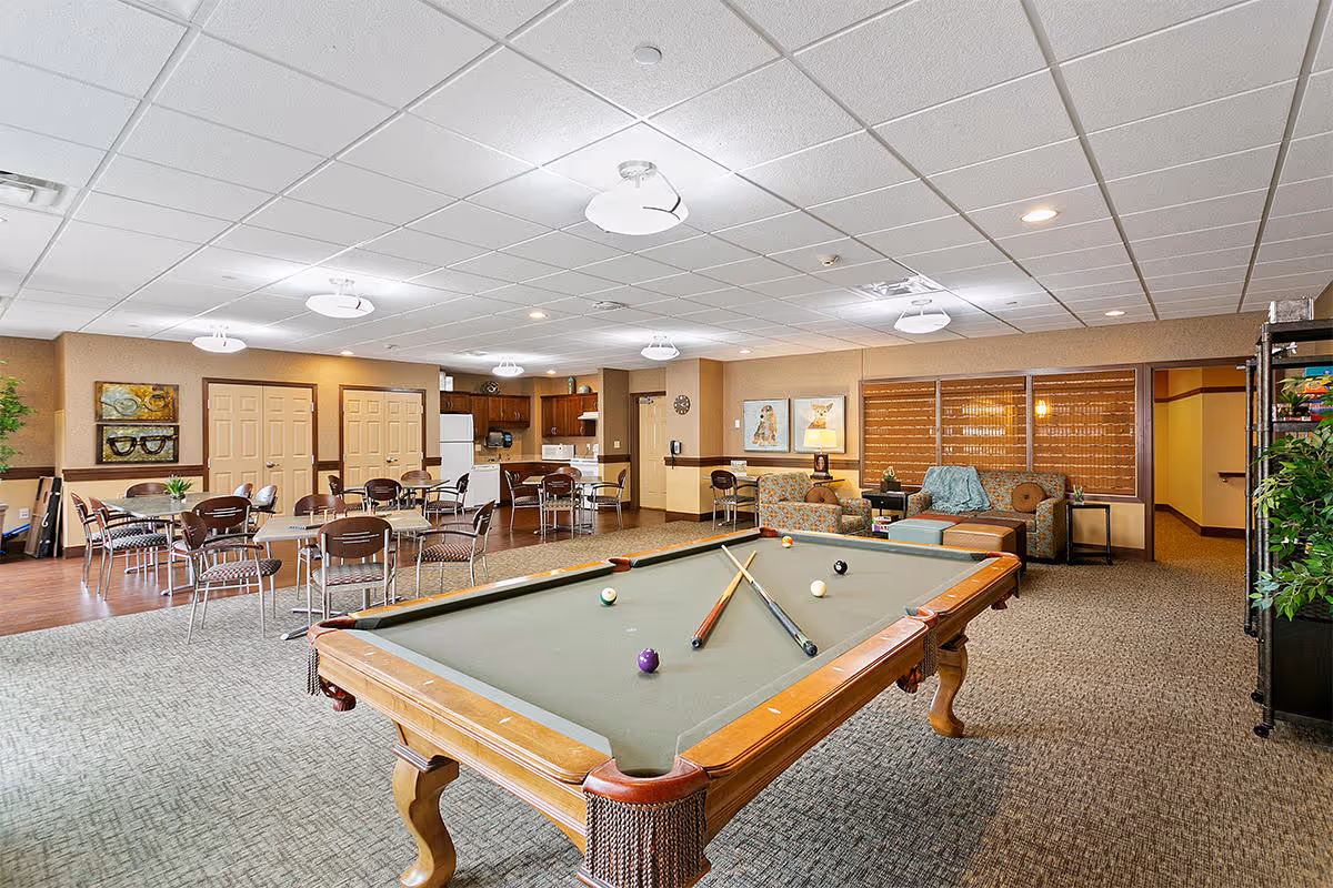 A spacious common area in a senior living facility featuring a pool table with cues and balls, several tables and chairs for dining or activities, a small kitchen area with appliances, and a cozy seating area with couches and artwork on the walls.