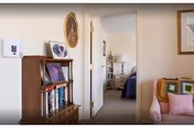 View of a cozy interior space showing a wooden bookshelf with books and framed pictures on the left, a doorway leading to a bedroom with a bed and lamp visible, and part of a pink armchair with a colorful crocheted blanket on the right.