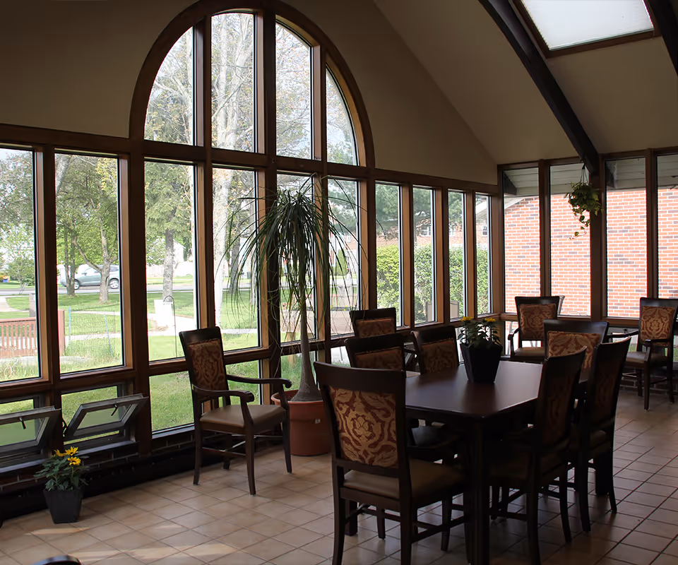 Sunlit dining area with a long wooden table, upholstered chairs, large arched windows overlooking greenery, and potted plants.