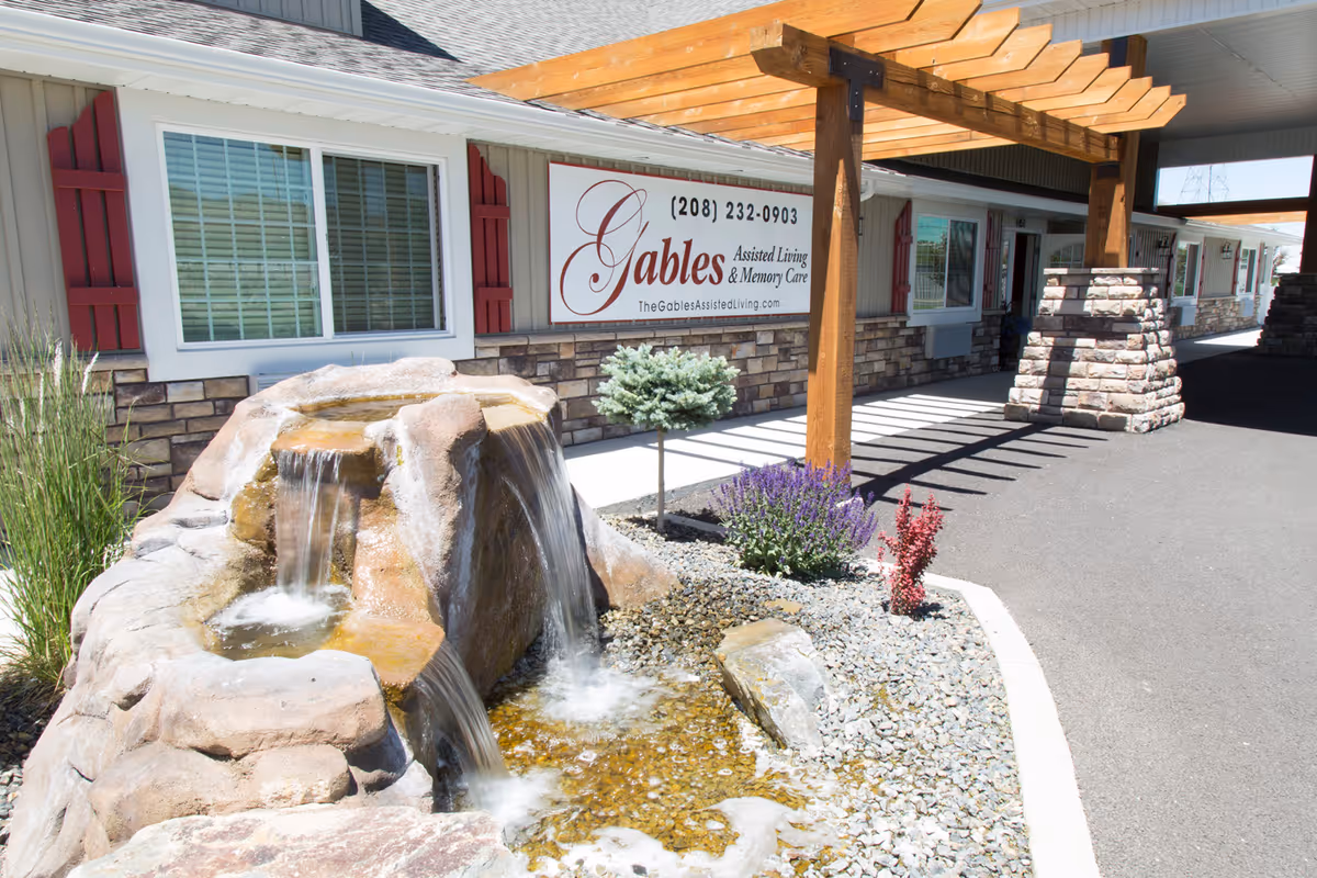 Outdoor view of The Gables Memory Care of Pocatello facility showing a stone water fountain with cascading water, a small landscaped area with plants and rocks, a wooden pergola structure, and the building exterior with windows and a sign displaying the facility name, phone number, and website.