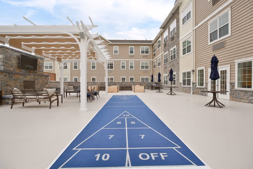 Outdoor courtyard area at Merrill Gardens at Glen Mills featuring a shuffleboard court in the center, seating areas with chairs and tables under a pergola, a stone fireplace with a mounted TV, and several tables with closed umbrellas along the building walls.