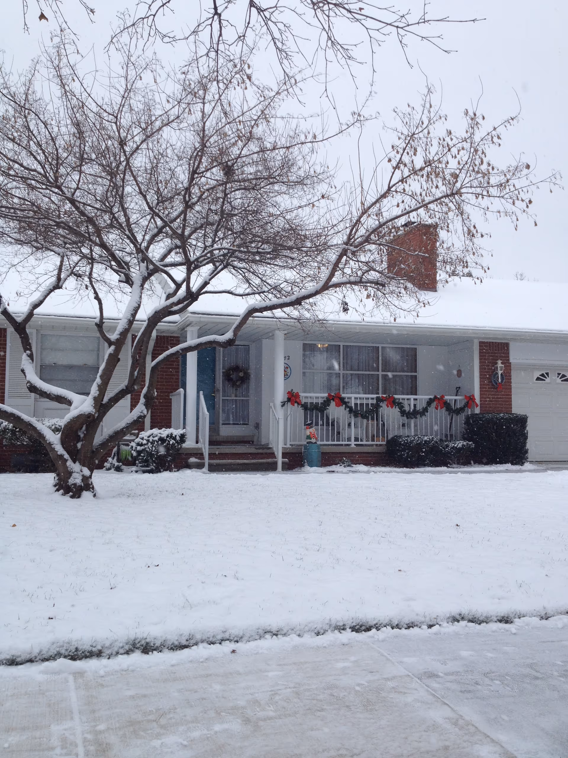A single-story brick house covered in snow with a leafless tree in the front yard. The porch is decorated with green garlands and red bows, and there is a wreath on the front door. Snow blankets the lawn and driveway.