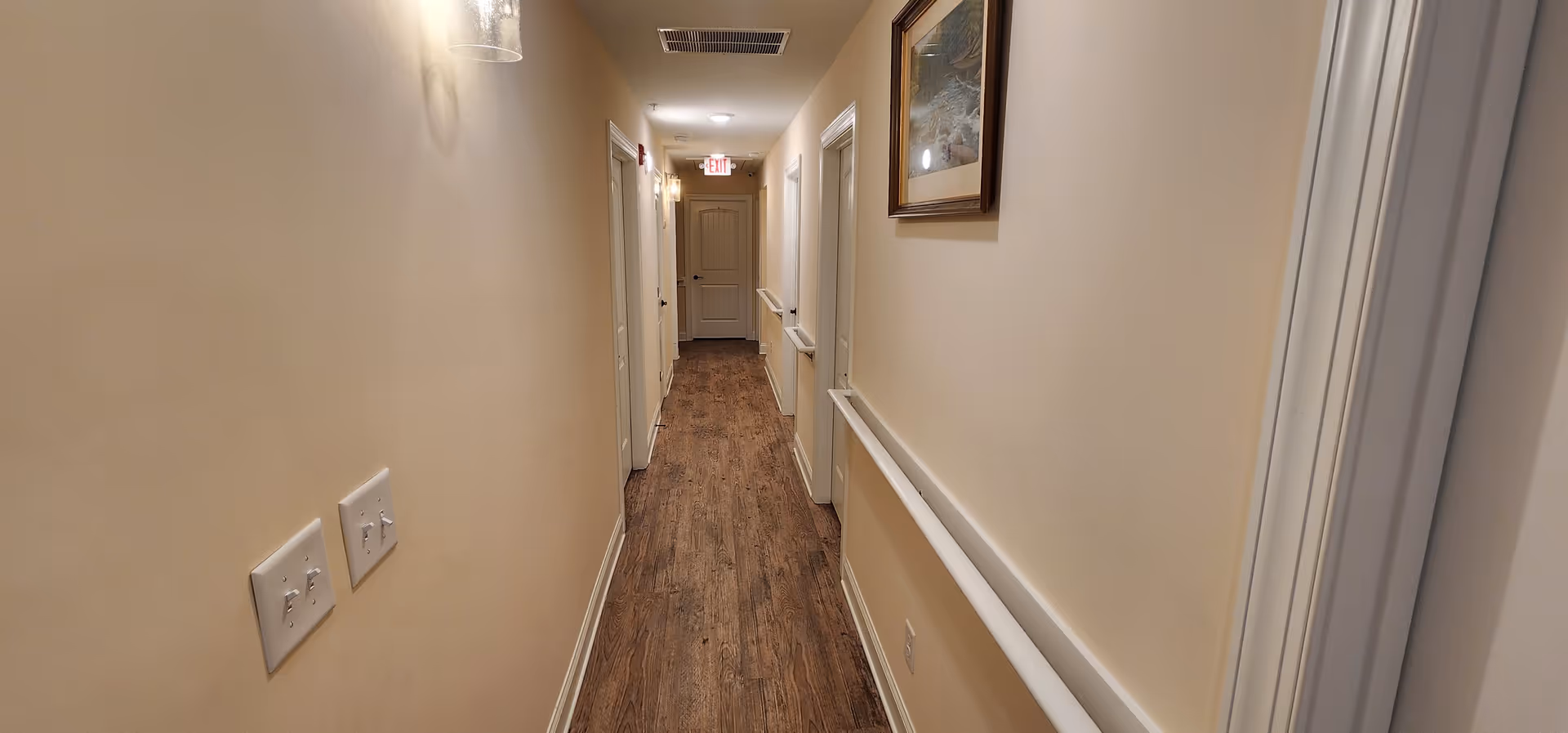A long, narrow hallway with wooden flooring and beige walls. There are several white doors on both sides of the hallway, handrails along the walls, light fixtures mounted on the walls, and an exit sign above a door at the end of the hallway. A framed picture is hanging on the right wall.