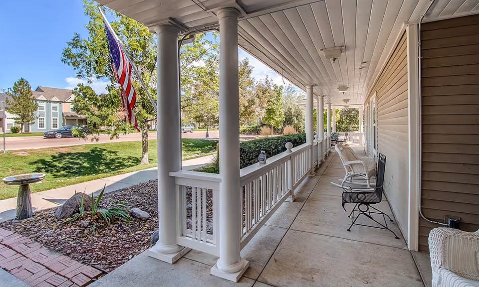 A covered porch area with white columns and railings, several chairs including wicker and metal ones, overlooking a landscaped garden with rocks, plants, and a birdbath. An American flag is mounted on one of the columns, and a street with houses and trees is visible in the background under a blue sky.