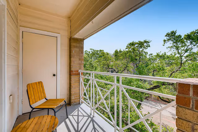 A small balcony with two wooden chairs and a white door, overlooking green trees and a clear blue sky.