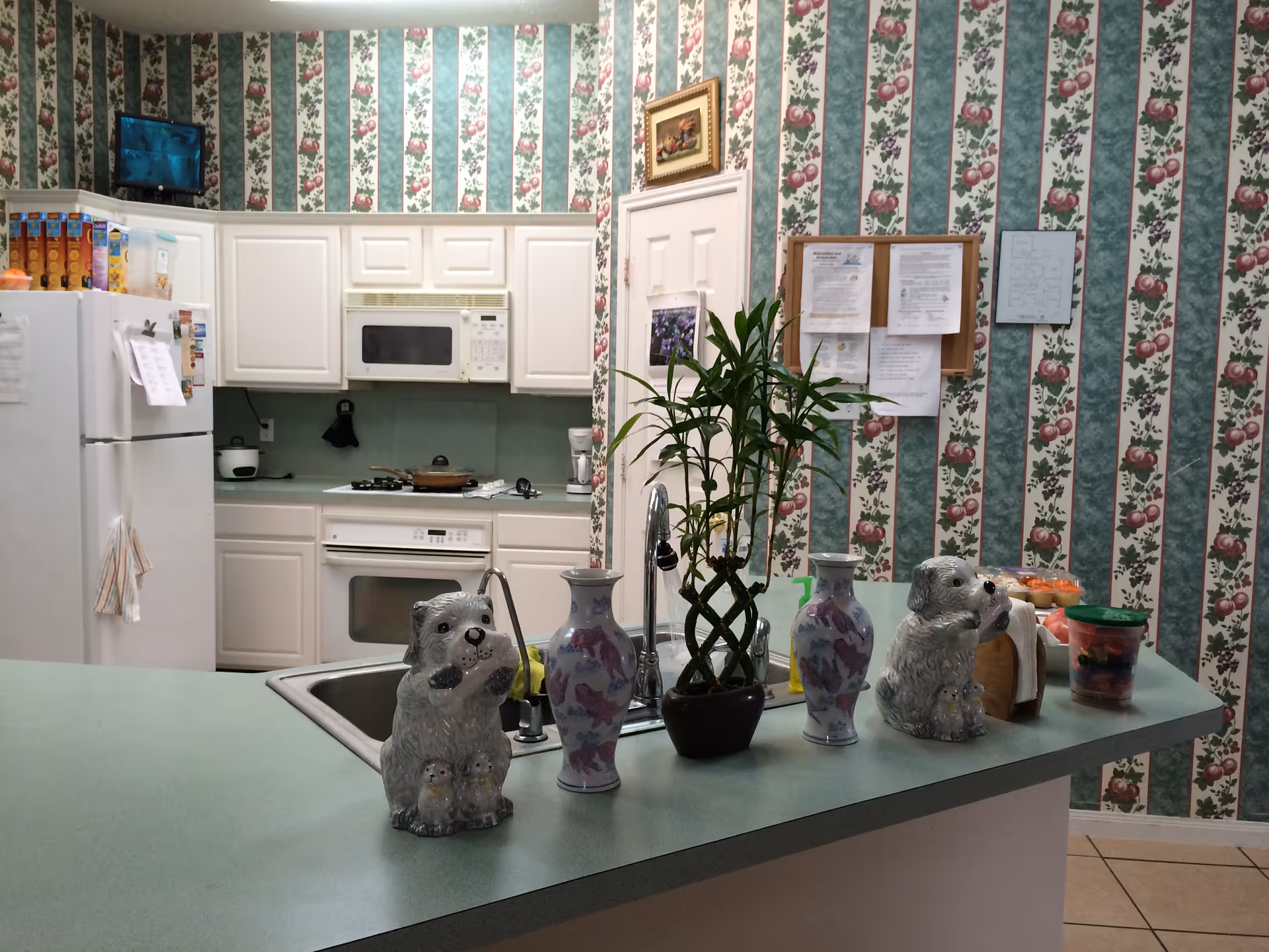 Kitchen with a green countertop island holding ceramic dog figurines, vases and a potted plant, white cabinets and appliances, and floral striped wallpaper.