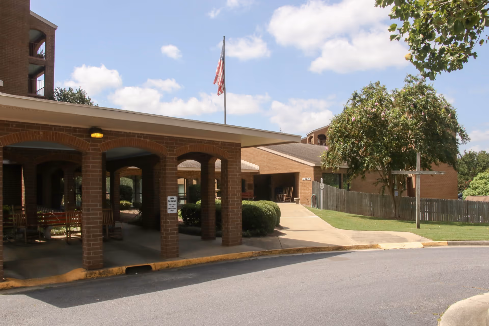 Exterior view of a senior living facility entrance with a covered drop-off area supported by brick columns. There is an American flag on a flagpole above the roof, a tree and bushes near the building, and a sidewalk leading to an office entrance. The sky is partly cloudy.