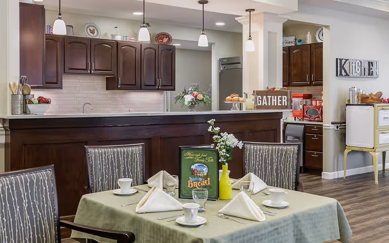 A dining area in a senior living facility with a table set for four, featuring white napkins, cups, and glasses. Behind the table is a kitchen counter with dark wood cabinets, a sink, and decorative items including a flower vase and a sign that says 'GATHER'. The room has warm lighting and a cozy atmosphere.
