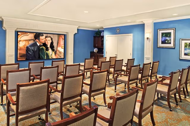 A small theater room with rows of wooden chairs facing a large screen displaying a scene from a movie. The room has blue walls, framed pictures, and a carpeted floor.