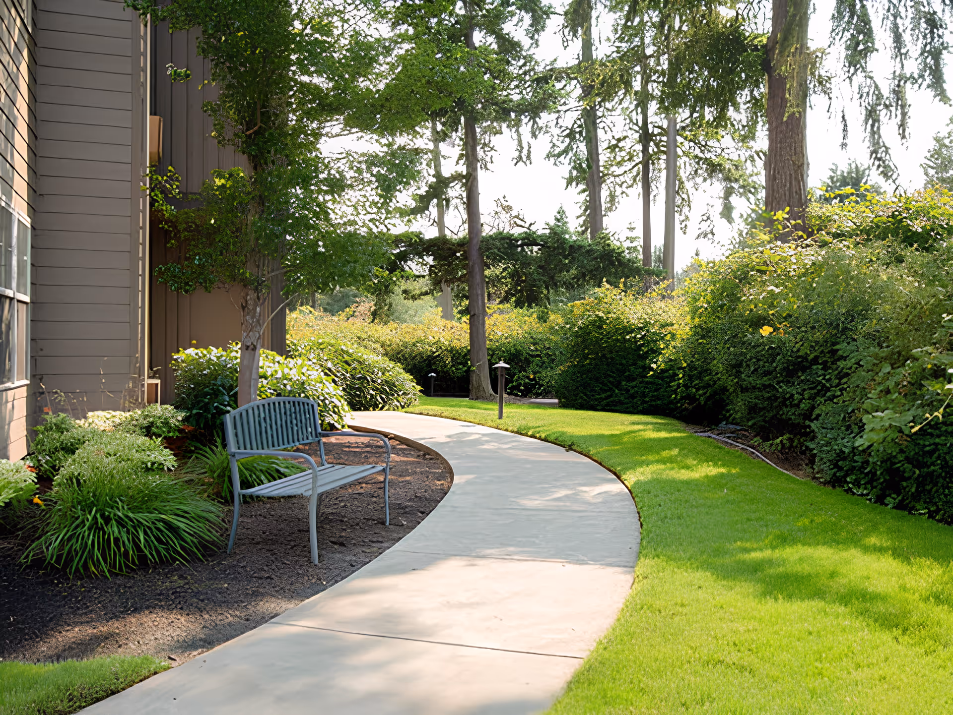 A curved concrete pathway in a garden area beside a building with a metal bench under a tree. The garden is lush with green grass, bushes, and tall trees in the background.
