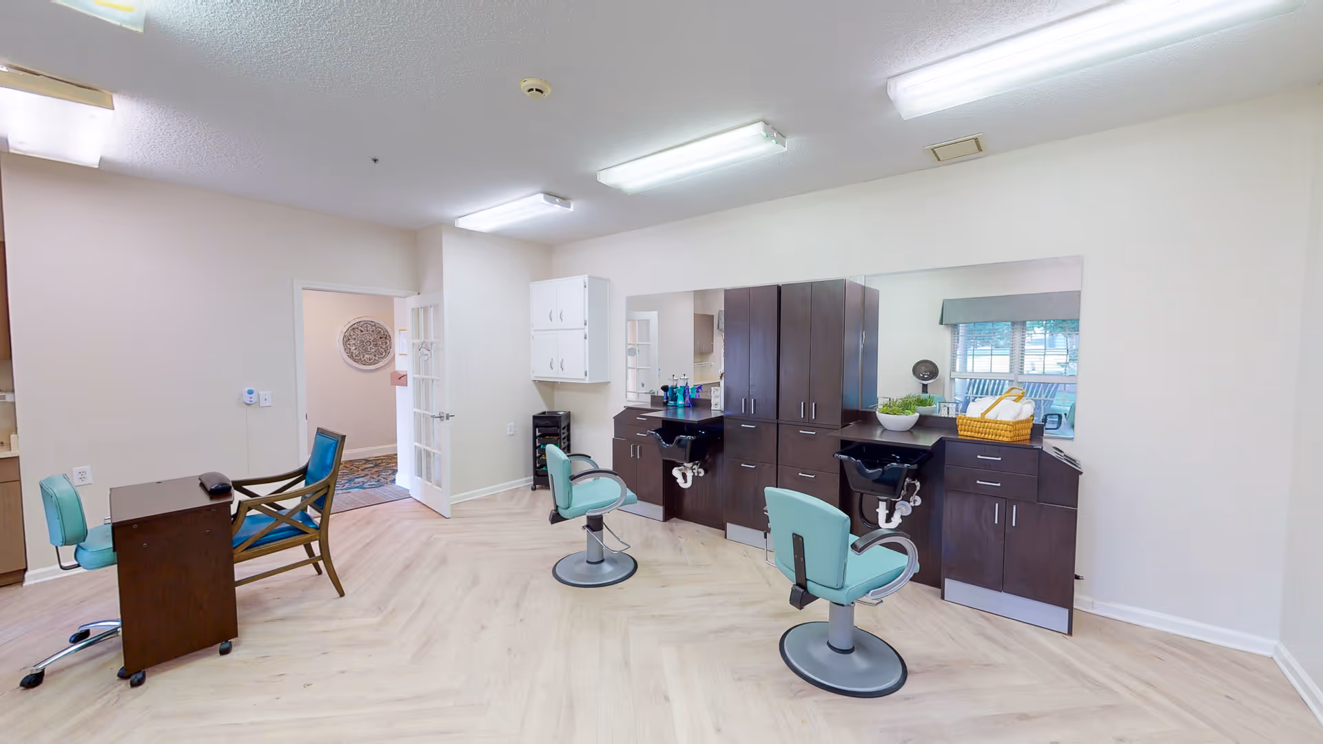 Interior view of a salon area in a senior living facility with two teal salon chairs facing black wash basins and dark wood cabinetry. There is a desk with two chairs on the left side, and a large mirror on the wall behind the salon stations. The room has light-colored wood flooring and white walls with fluorescent ceiling lights.