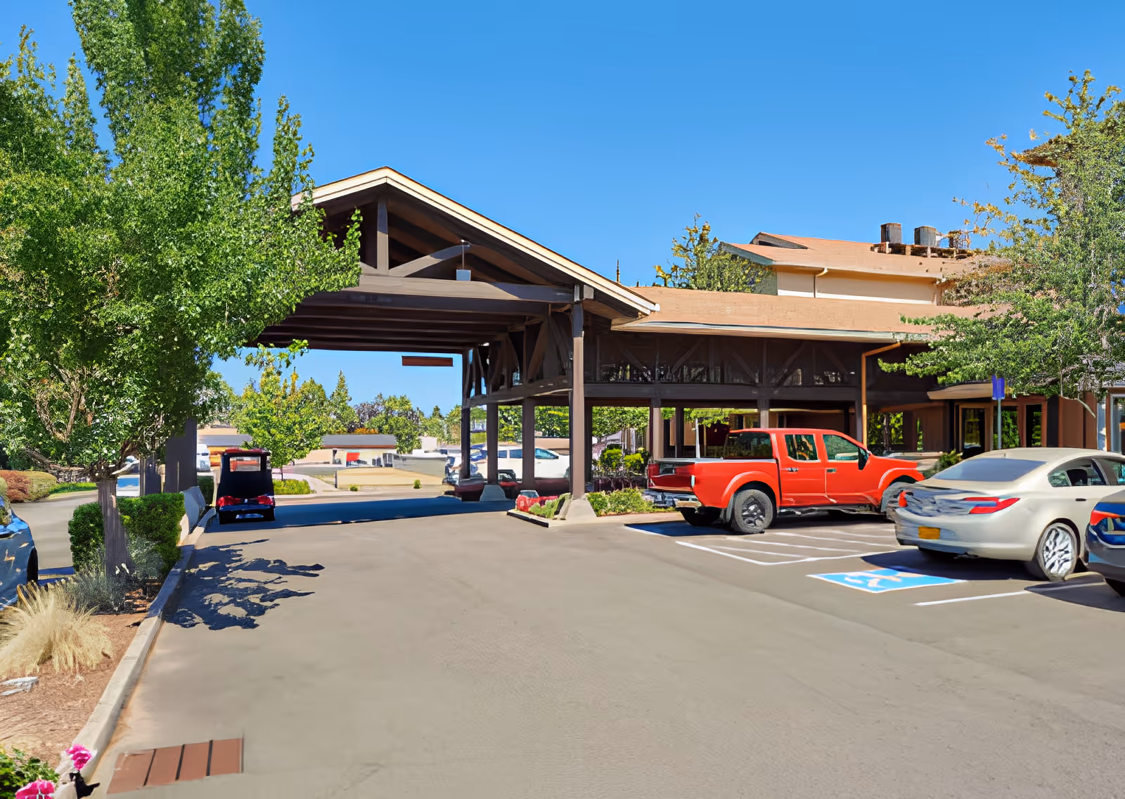 Exterior view of Elliott Residence showing a covered entrance with a wooden roof structure, several parked cars including a red pickup truck, and surrounding greenery under a clear blue sky.
