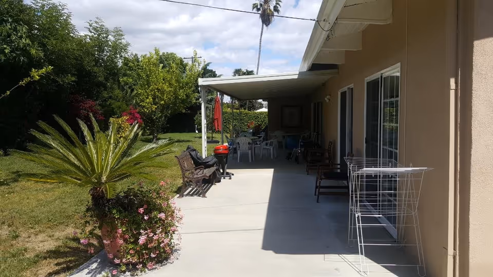 Covered patio with outdoor chairs and tables beside a lawn and potted plants.