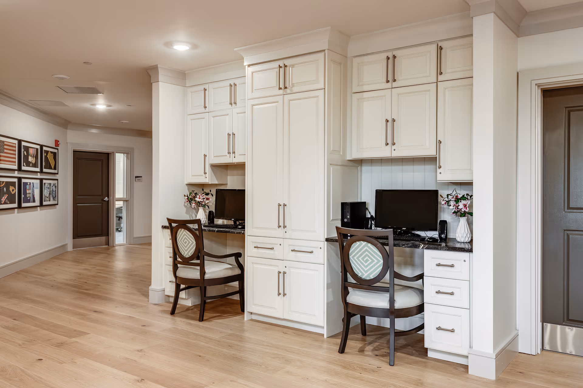 A hallway area in a senior living facility featuring two built-in white desks with dark granite countertops, each equipped with a computer and a wooden chair with a patterned cushion. There are white cabinets above and below the desks, and vases with flowers on each desk. The hallway has light wood flooring, framed pictures on the wall, and a closed dark brown door at the end.