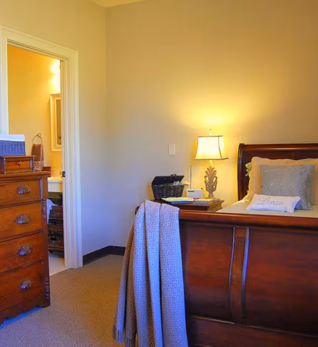 A cozy bedroom with a wooden bed frame and a bedside table with a lamp. The bed has pillows, including one with the word 'Dream' on it. A light-colored blanket is draped over the footboard. To the left, there is a wooden dresser and an open door leading to a bathroom with a sink and mirror visible.