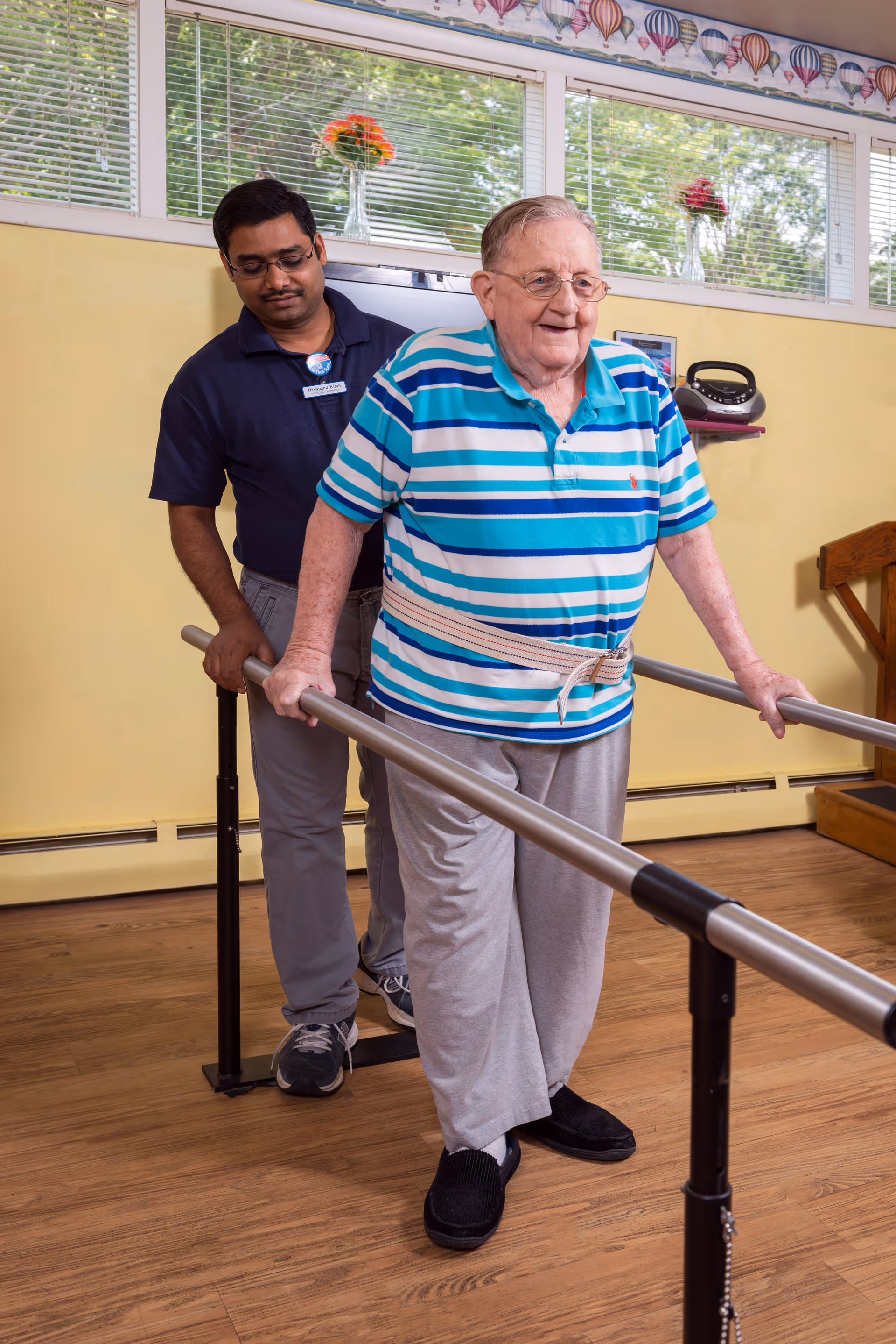 An elderly man wearing a blue and white striped shirt and gray pants is walking with the support of parallel bars in a room with wooden flooring. A caregiver stands behind him, providing assistance. The room has yellow walls, large windows with blinds, and flower vases on the windowsill.