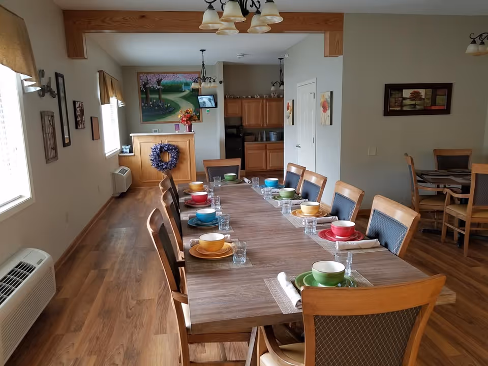 Communal dining room with a long wooden table set with colorful bowls and glasses, surrounded by chairs and an open kitchen in the background.
