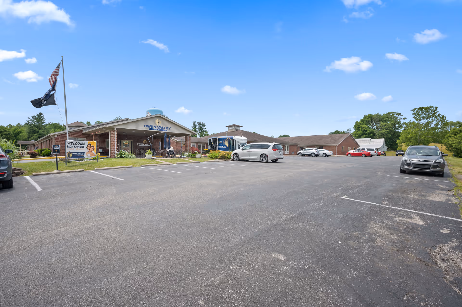 Exterior view of Owen Valley Rehabilitation and Healthcare Center showing a large parking lot with several parked cars, a flagpole with American and POW flags, and a sign welcoming back families. The building is a single-story brick structure with a covered entrance and a water tower in the background under a blue sky with scattered clouds.