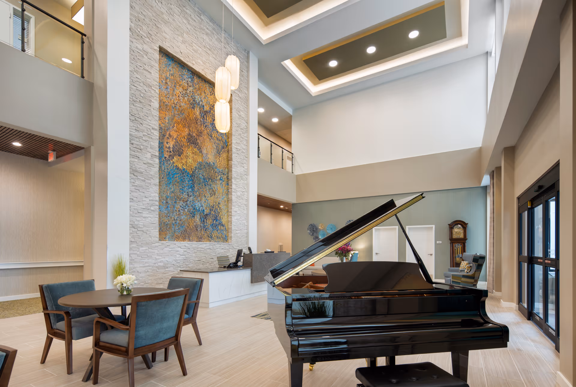 Spacious and modern senior living facility lobby with a black grand piano in the foreground, a round table with four chairs, a reception desk, tall textured wall art, and high ceilings with recessed lighting.
