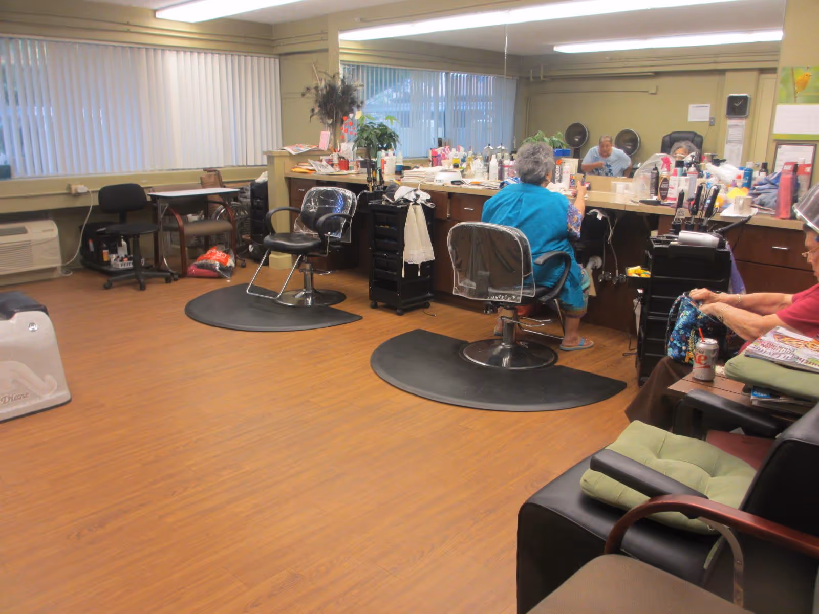 Interior of a salon area in a senior living facility with two elderly women seated in salon chairs facing a large mirror. The room has wooden flooring, salon equipment, and various hair care products on the counter. There are additional chairs and a window with vertical blinds in the background.