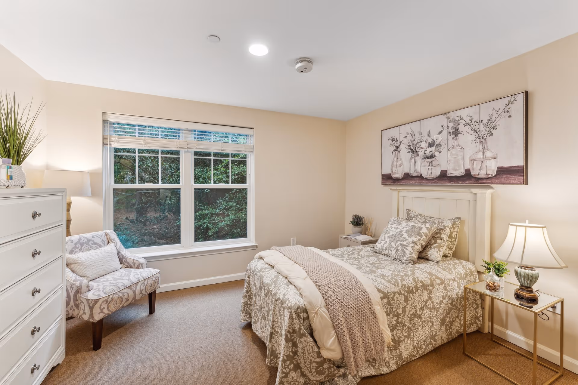 Well-lit bedroom with a single bed dressed in patterned bedding, an armchair by a large window, dresser, and bedside tables with lamps.