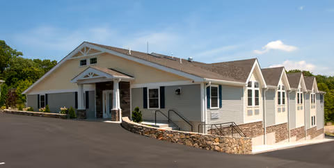 Exterior view of a single-story assisted living facility building with beige siding, stone accents, and a gabled roof under a clear blue sky. The building has multiple windows and a main entrance with a small covered porch. There is a paved driveway and a stone retaining wall in front of the building.