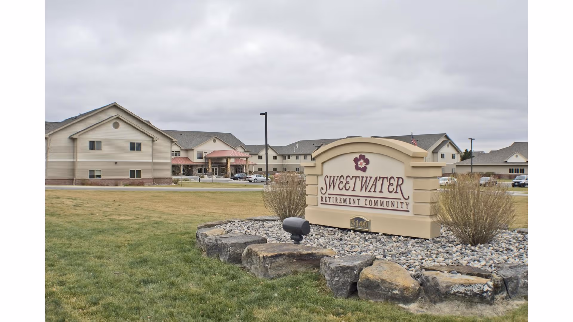 View of the Sweetwater Retirement Community sign in front of a large, multi-building residential facility with beige siding and a gray roof under a cloudy sky. The sign is surrounded by rocks and small bushes, with a grassy lawn in the foreground.