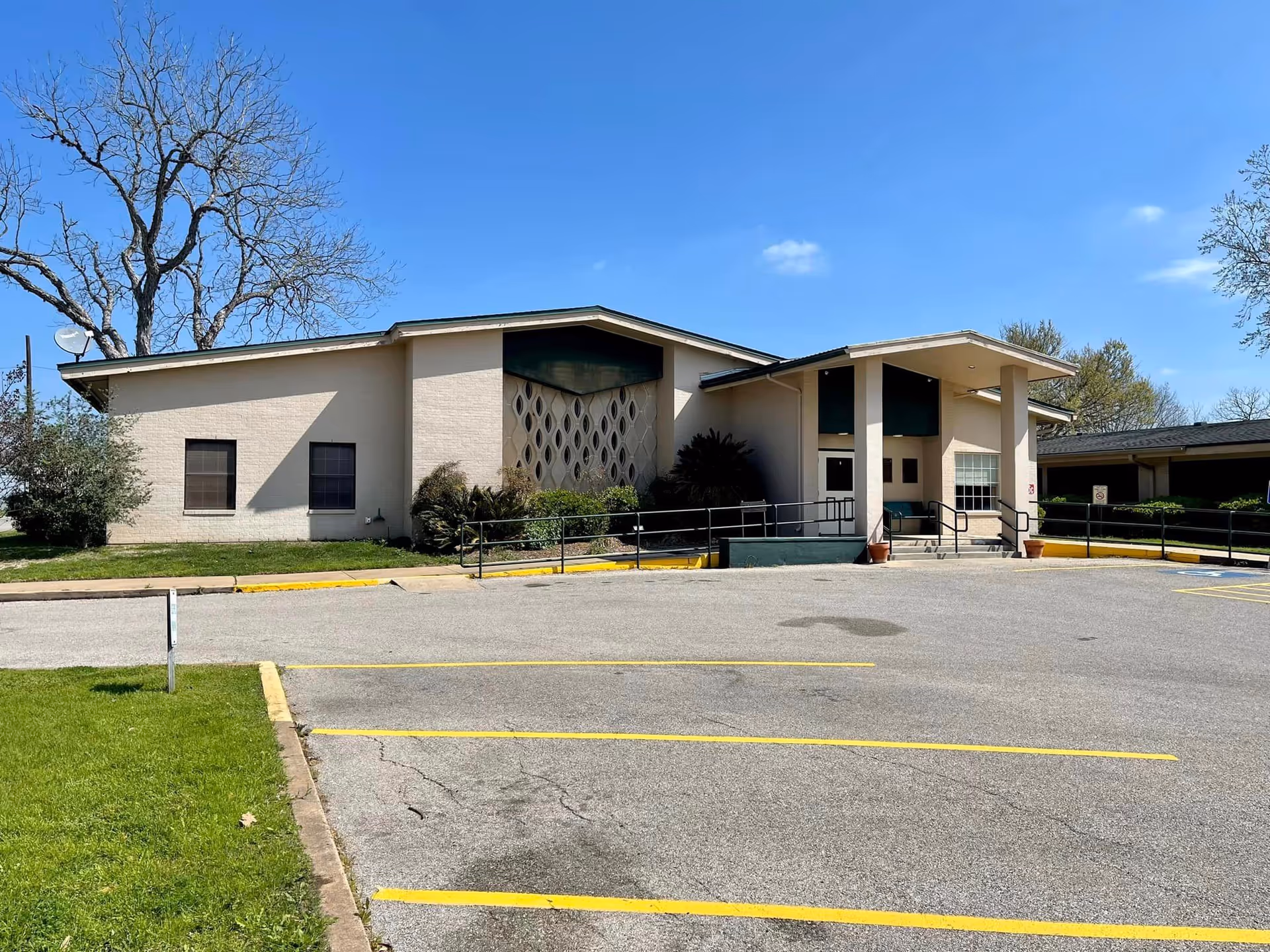 Exterior view of Shiner Nursing & Rehab Center building with a beige facade, a ramp leading to the entrance, some bushes and trees around, and an empty parking lot with yellow lines under a clear blue sky.