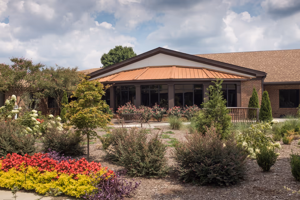 Front entrance of a single-story senior living building with a copper-colored awning and landscaped flower beds under a partly cloudy sky.