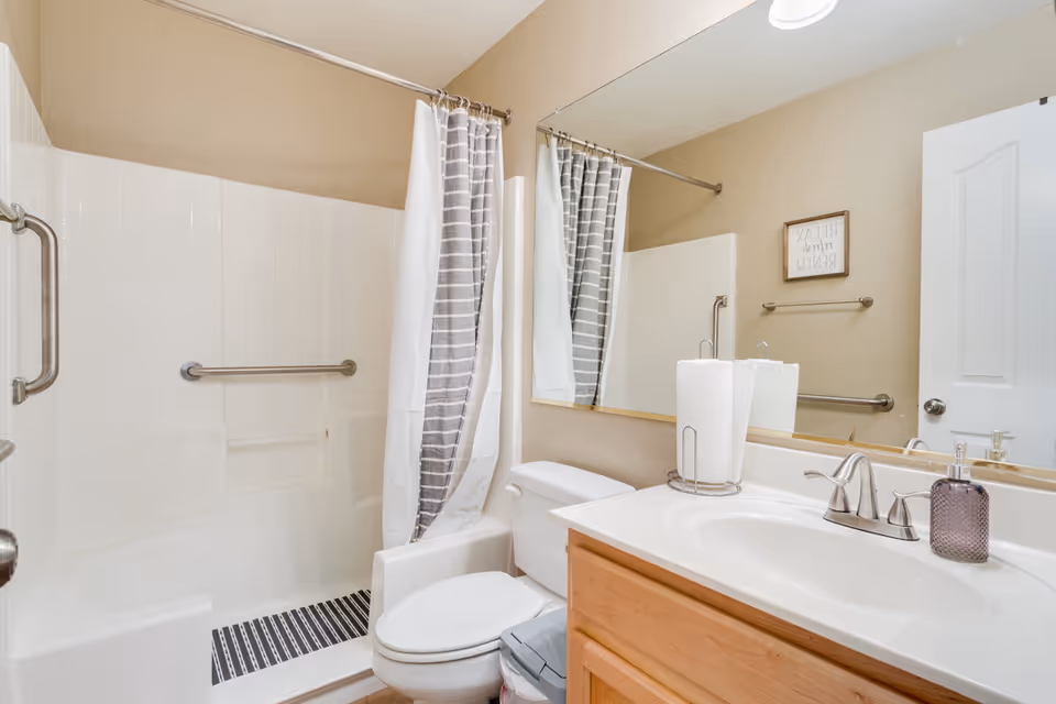 A clean bathroom featuring a white toilet, a wooden vanity with a white countertop and sink, a large mirror above the sink, and a shower area with white tiled walls and a gray and white striped shower curtain. There are metal grab bars installed near the shower and toilet for accessibility. A small framed sign is visible on the beige wall above the towel rack.