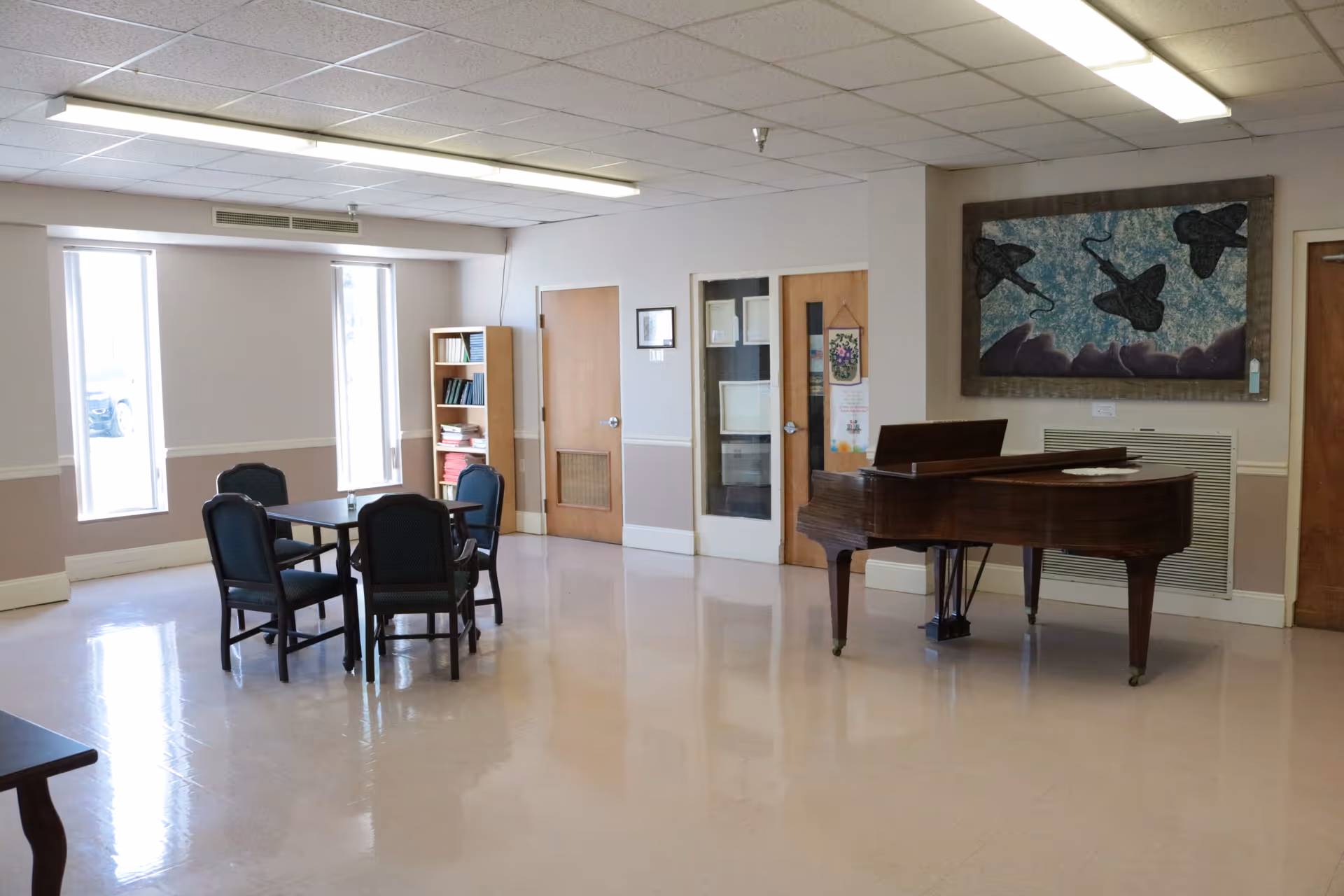 A spacious room with a polished floor featuring a wooden grand piano on the right side and a square table with four chairs on the left. There are two tall windows letting in natural light, a wooden bookshelf filled with books, and three wooden doors along the back wall. A framed artwork depicting three black birds flying against a blue sky hangs above the piano.