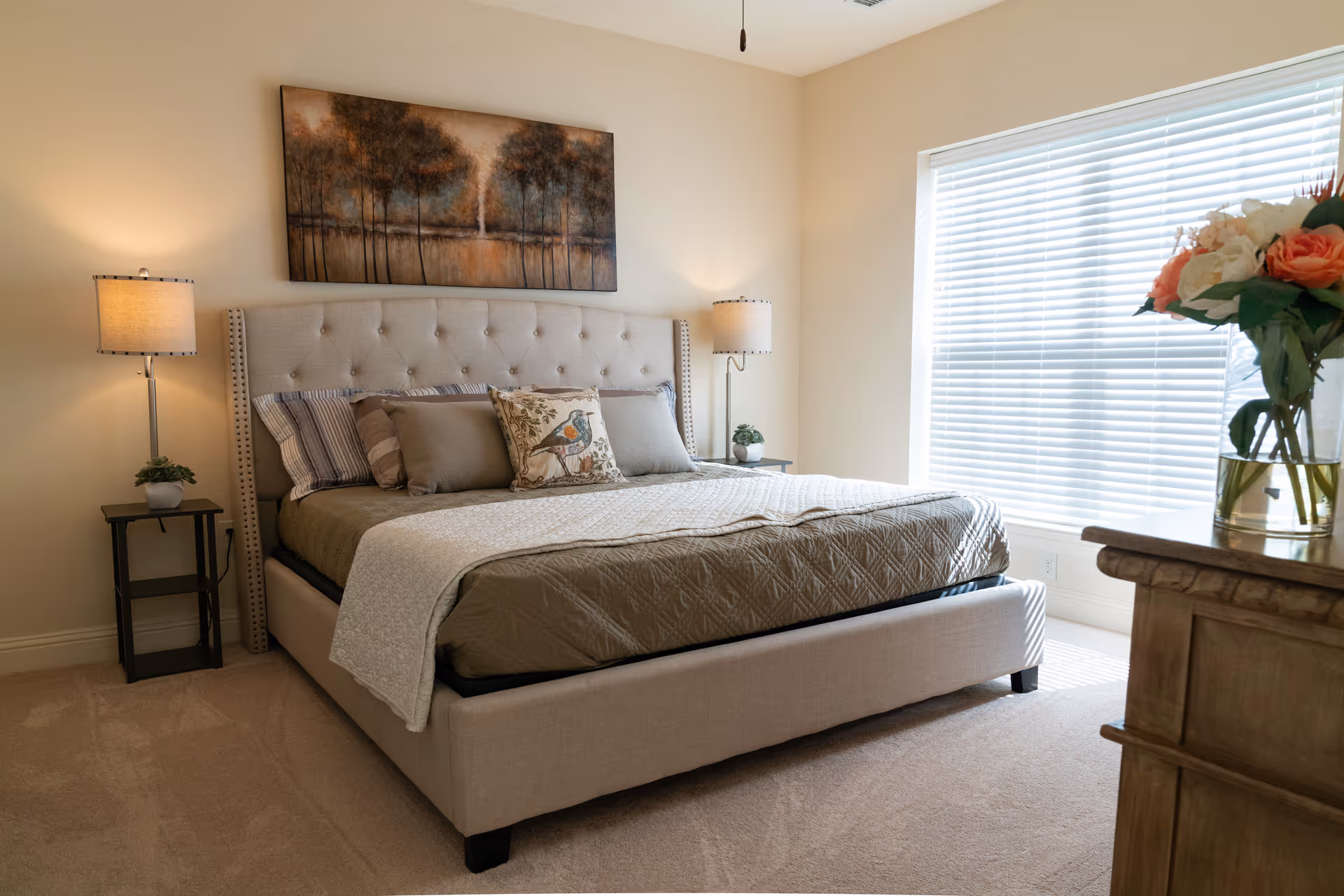 A neatly made bed with a beige upholstered headboard and multiple pillows, including one with a bird design. There are two bedside tables with lamps and small plants on each side of the bed. A large window with blinds allows natural light to fill the room. A wooden dresser with a vase of flowers is visible in the foreground. A painting of trees hangs above the bed.