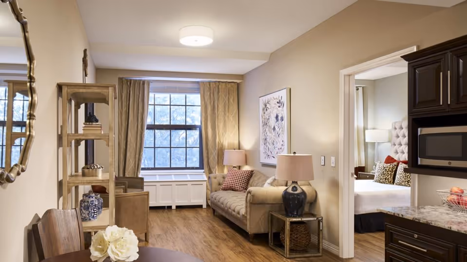 A cozy living room area in a senior living facility featuring a beige sofa with patterned cushions, a side table with a lamp, a wooden shelving unit with decorative items, and a large window with curtains. Adjacent to the living room is a partial view of a bedroom with a bed, pillows, and a bedside lamp. The kitchen area with dark cabinetry and a microwave is also partially visible.
