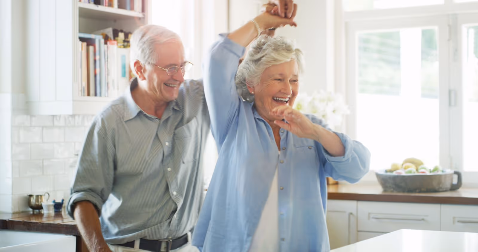 An elderly couple joyfully dancing together in a bright kitchen with white cabinets and a bowl of fruit on the counter in the background.
