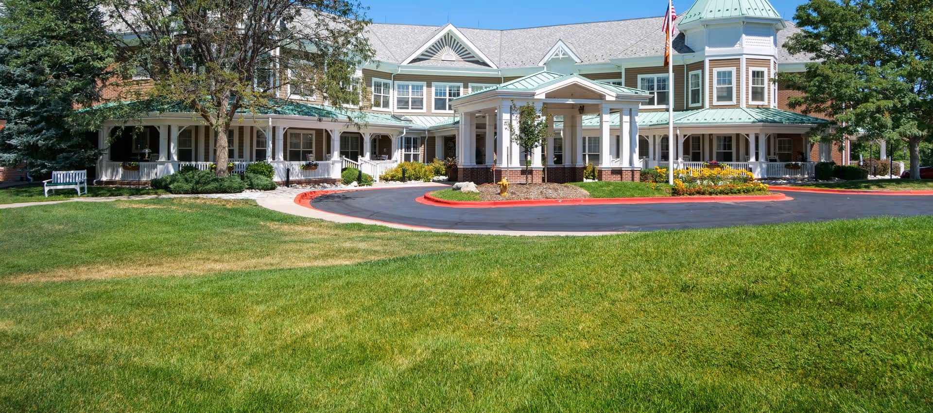 Front exterior view of a senior living facility with a circular driveway, green lawn, trees, and a large building featuring white columns, multiple windows, and a covered entrance with a flagpole displaying the American flag.