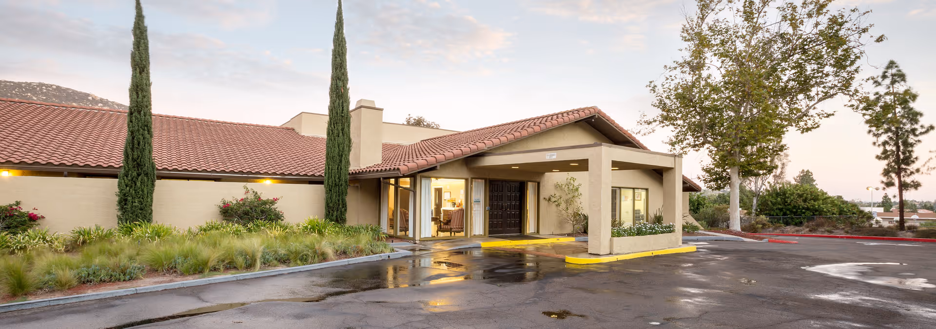 Front entrance of a single-story senior living facility with a covered porte-cochere, tile roof, landscaped beds, and a wet driveway at dusk.
