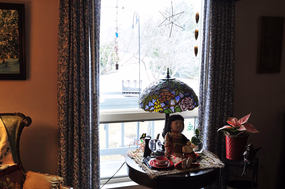 A cozy corner of a living room with a round wooden table in front of a window. On the table, there is a stained glass lamp with floral patterns, a doll dressed in traditional clothing, a glass, a red cup and saucer, and some small decorative items. The window is framed by patterned curtains, and there are hanging decorations visible outside the window. To the right of the table, there is a red potted plant on a small stand, and to the left, part of an upholstered armchair is visible.