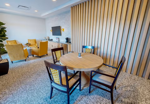 A cozy interior room with a round wooden table surrounded by four chairs with patterned cushions. In the background, there are four mustard-yellow armchairs arranged around a small table. A wall-mounted TV and a sideboard with decorative items and a coffee maker are visible. The room features a textured carpet and a wall with vertical wooden slats.