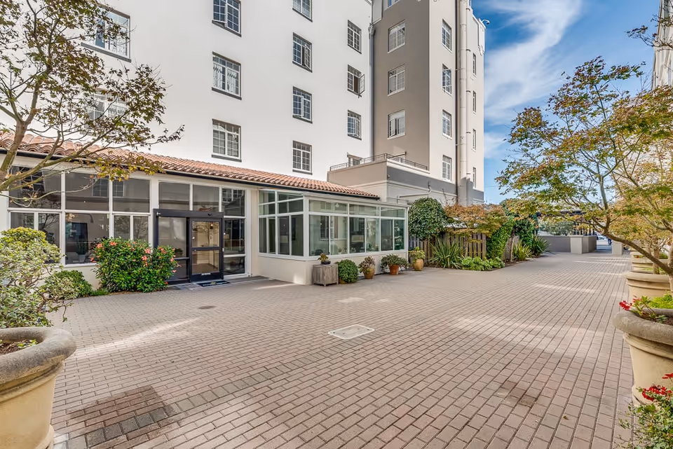 Outdoor courtyard area of a senior living facility with paved brick flooring, potted plants, small trees, and a multi-story building with large windows in the background under a partly cloudy sky.