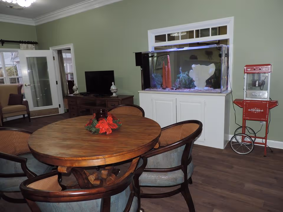 A cozy common area with a round wooden table surrounded by four cushioned chairs. On the table is a small holiday-themed centerpiece with red flowers and greenery. Against the wall is a large fish tank with colorful decorations inside, placed on a white cabinet. To the right of the fish tank is a vintage-style red popcorn machine on wheels. In the background, there is a wooden cabinet with a flat-screen TV and decorative items, and a glass door leading to another room with a beige armchair visible.