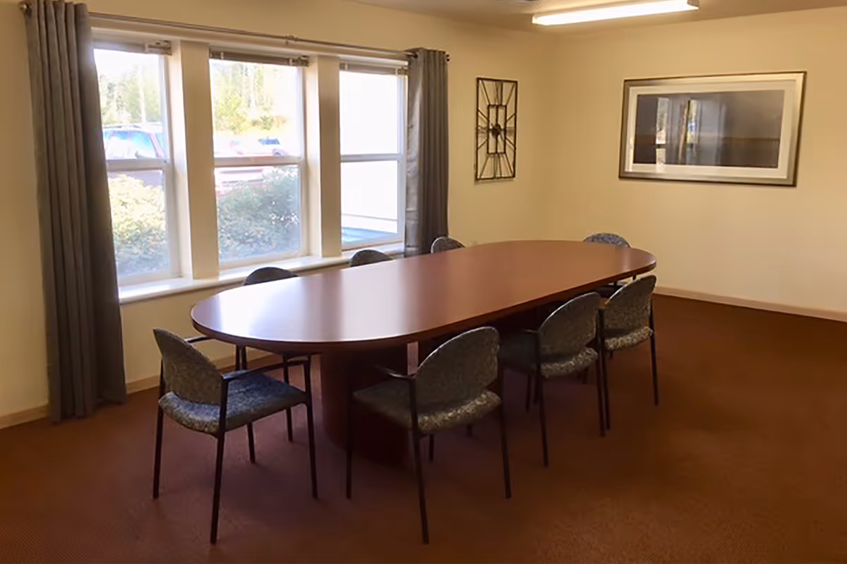 A conference room with a long oval wooden table surrounded by eight padded chairs. The room has beige walls, a brown carpet, three large windows with gray curtains, a framed picture on one wall, and a decorative wall clock.