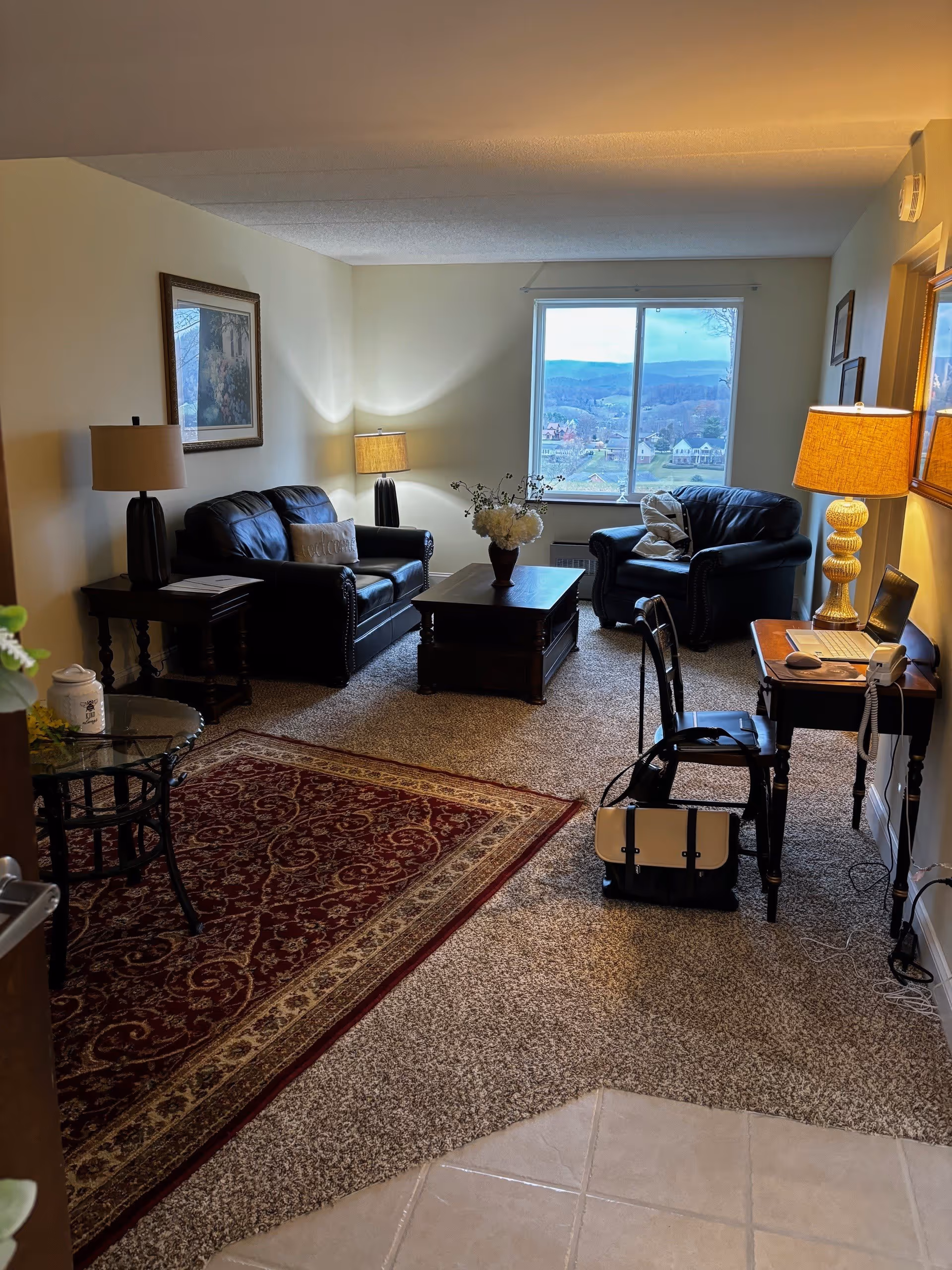 A cozy living room with two black leather armchairs and a matching loveseat arranged around a dark wooden coffee table with a vase of white flowers. There are two table lamps with beige shades on either side of the seating area, a framed picture on the wall, and a window showing a scenic view of hills and houses outside. A small wooden desk with a laptop, phone, and a chair is positioned near the right wall. The floor is carpeted with a patterned red area rug near the entrance.