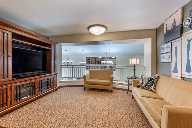 A cozy living room area with a large wooden entertainment center holding a flat-screen TV on the left. There is a beige sofa on the right with patterned pillows and a small side table with a lamp. In the background, a smaller beige loveseat is positioned near a railing overlooking a lower level with hanging chandeliers.
