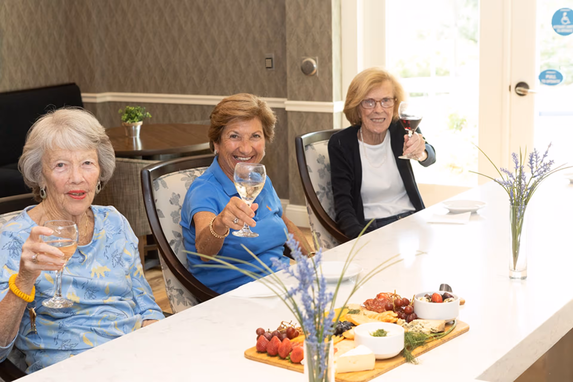 Three elderly women sitting at a white counter in a well-lit room, each holding a glass of wine and smiling towards the camera. In front of them on the counter is a wooden board with assorted fruits, cheeses, and dips, along with a vase containing lavender flowers.