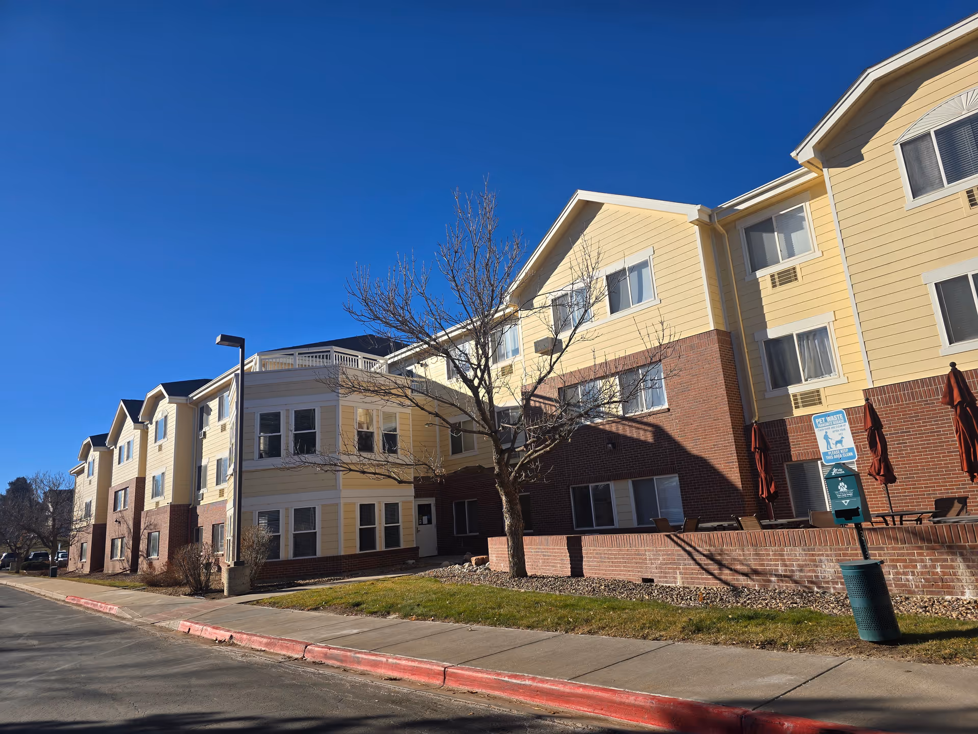 Three-story yellow-and-brick assisted living building with a sidewalk, leafless tree, and outdoor seating under a clear blue sky.
